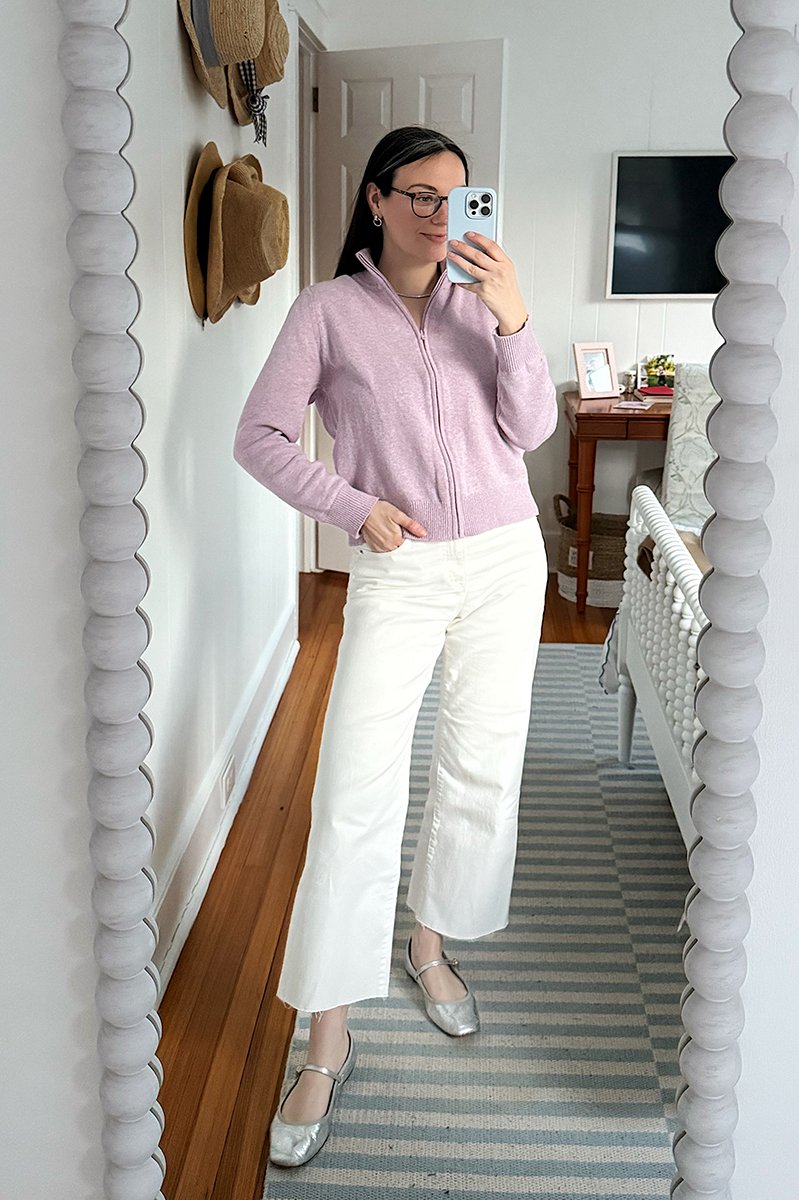 Carly Riordan taking a mirror selfie wearing a lavender zip up and white jeans. 