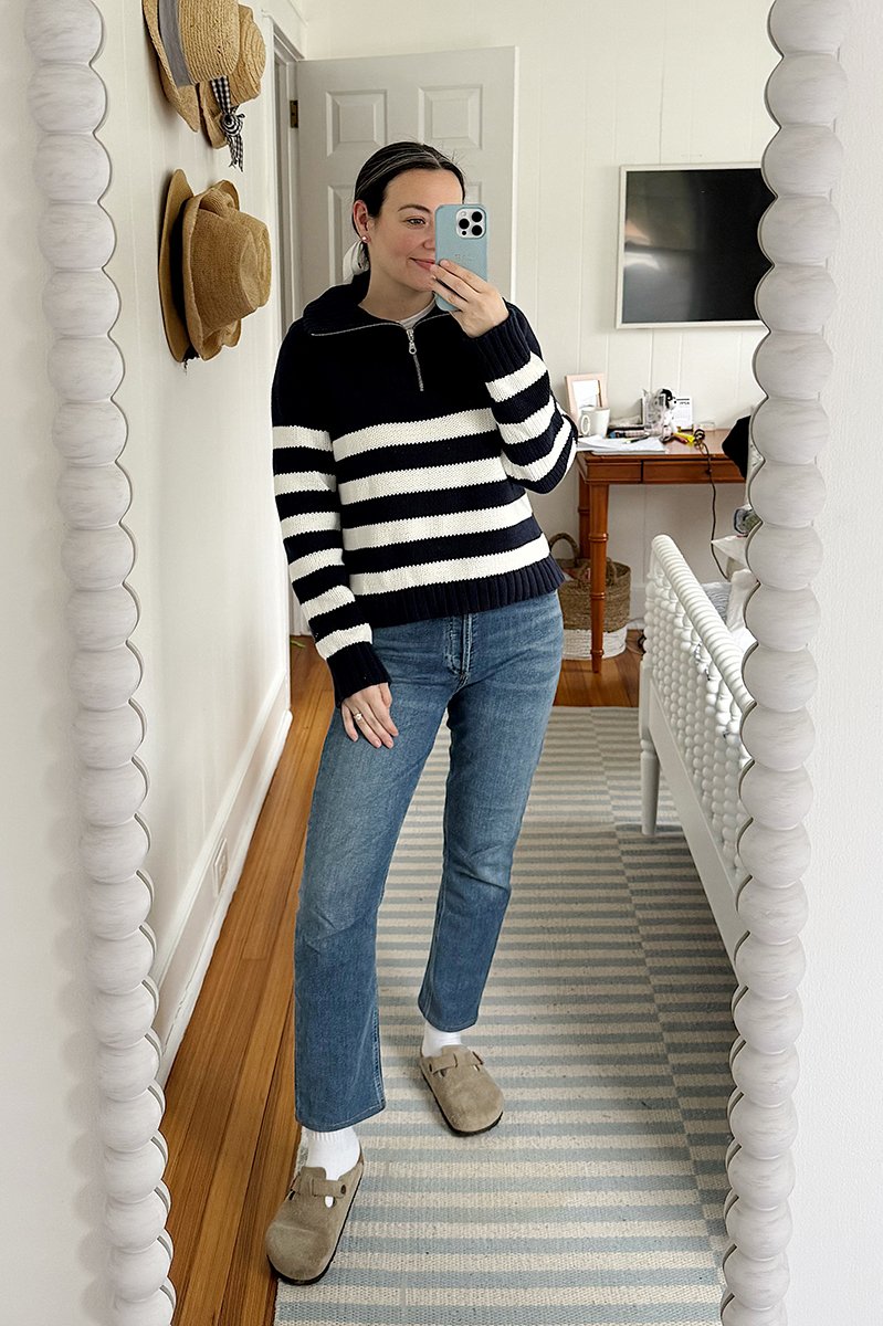 Carly Riordan taking a mirror selfie wearing a navy and white striped sweater and clogs.